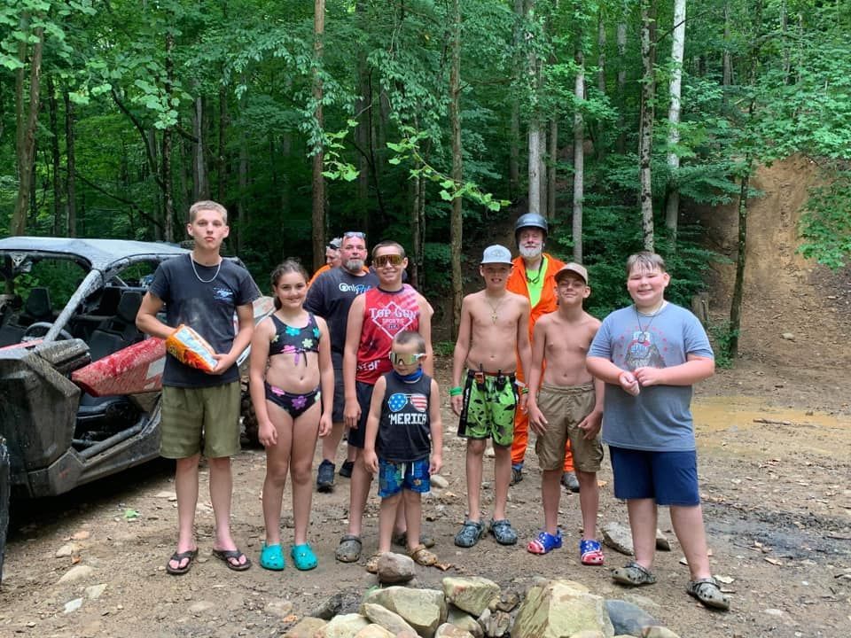 Group of people standing in a wooded area near a UTV. Some children are shirtless. The group smiles and looks at the camera.