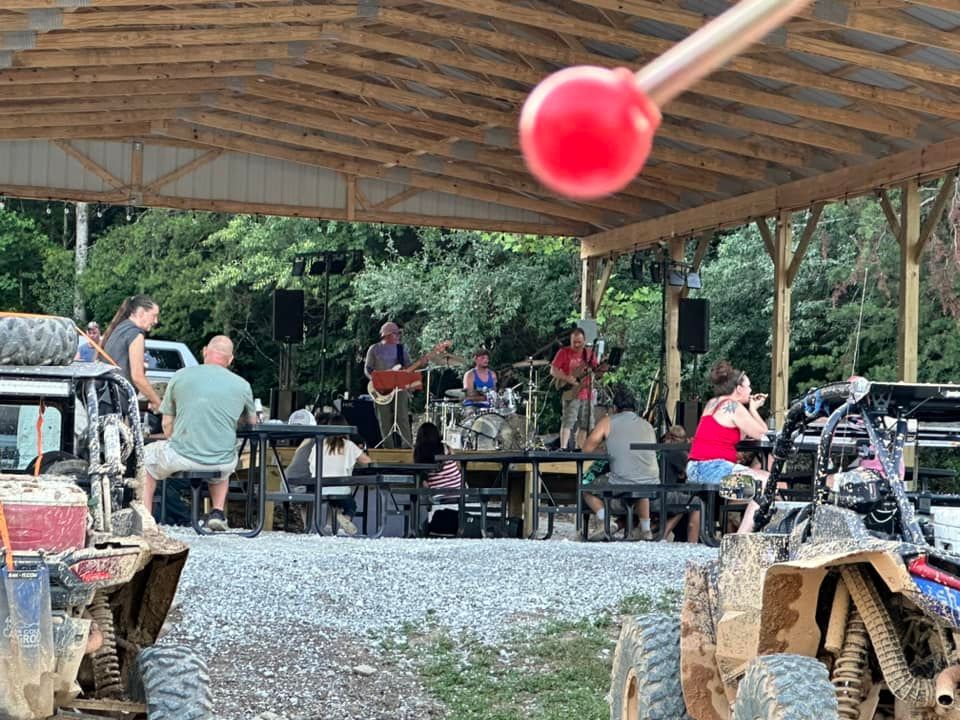 Band playing under a pavilion for people gathered outdoors, some seated at tables. Red mallet in foreground.