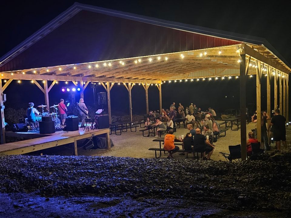 Band plays under a covered outdoor stage, with seated audience at picnic tables, at night.