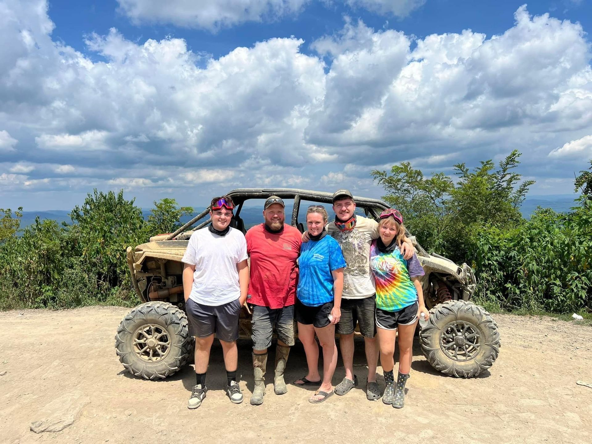 Group of five people posing with a dirty off-road vehicle on a mountain path under a cloudy sky.