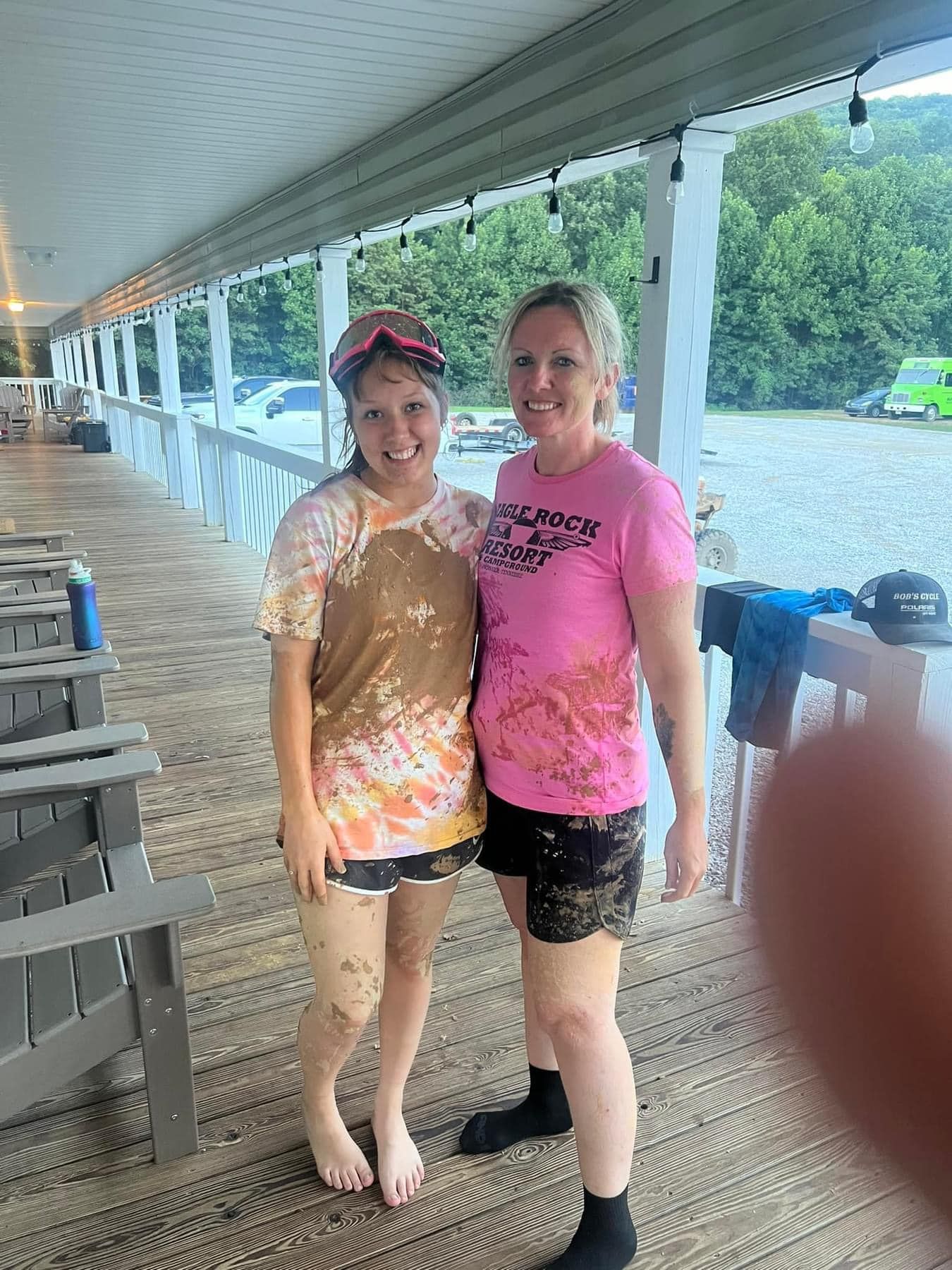 Two women covered in mud stand on a porch. One wears a pink shirt, the other a tie-dye shirt.