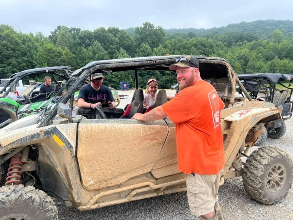 People near a muddy side-by-side vehicle. Man in orange shirt smiles. Others sit inside. Mountain backdrop.