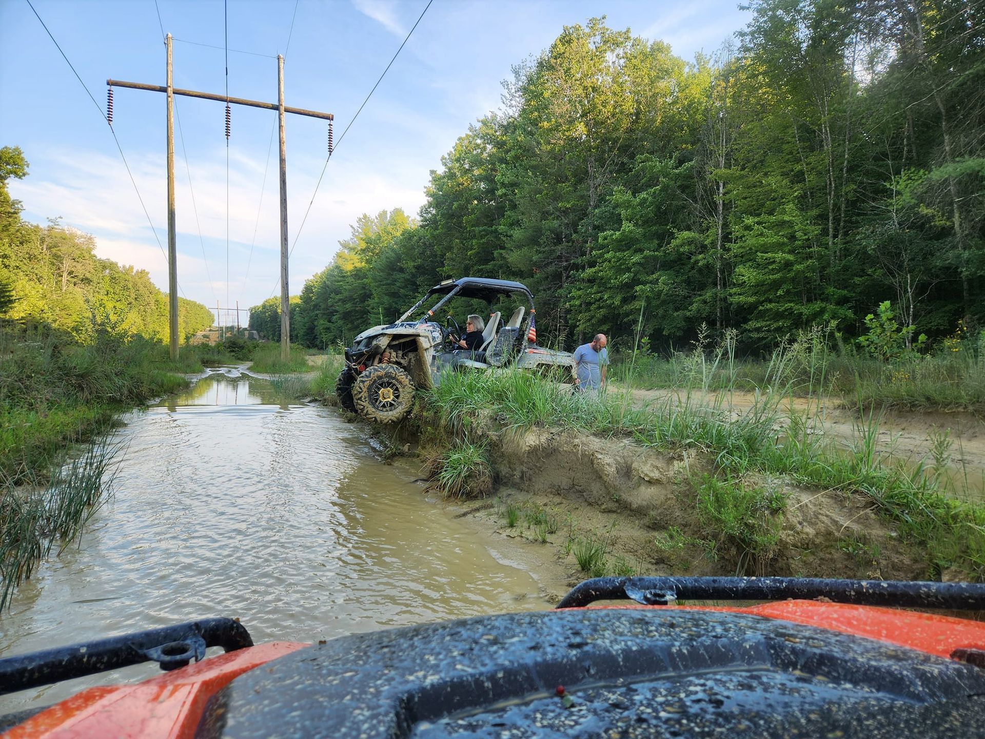 ATV stuck in muddy water with a person standing nearby, power lines and trees in the background.