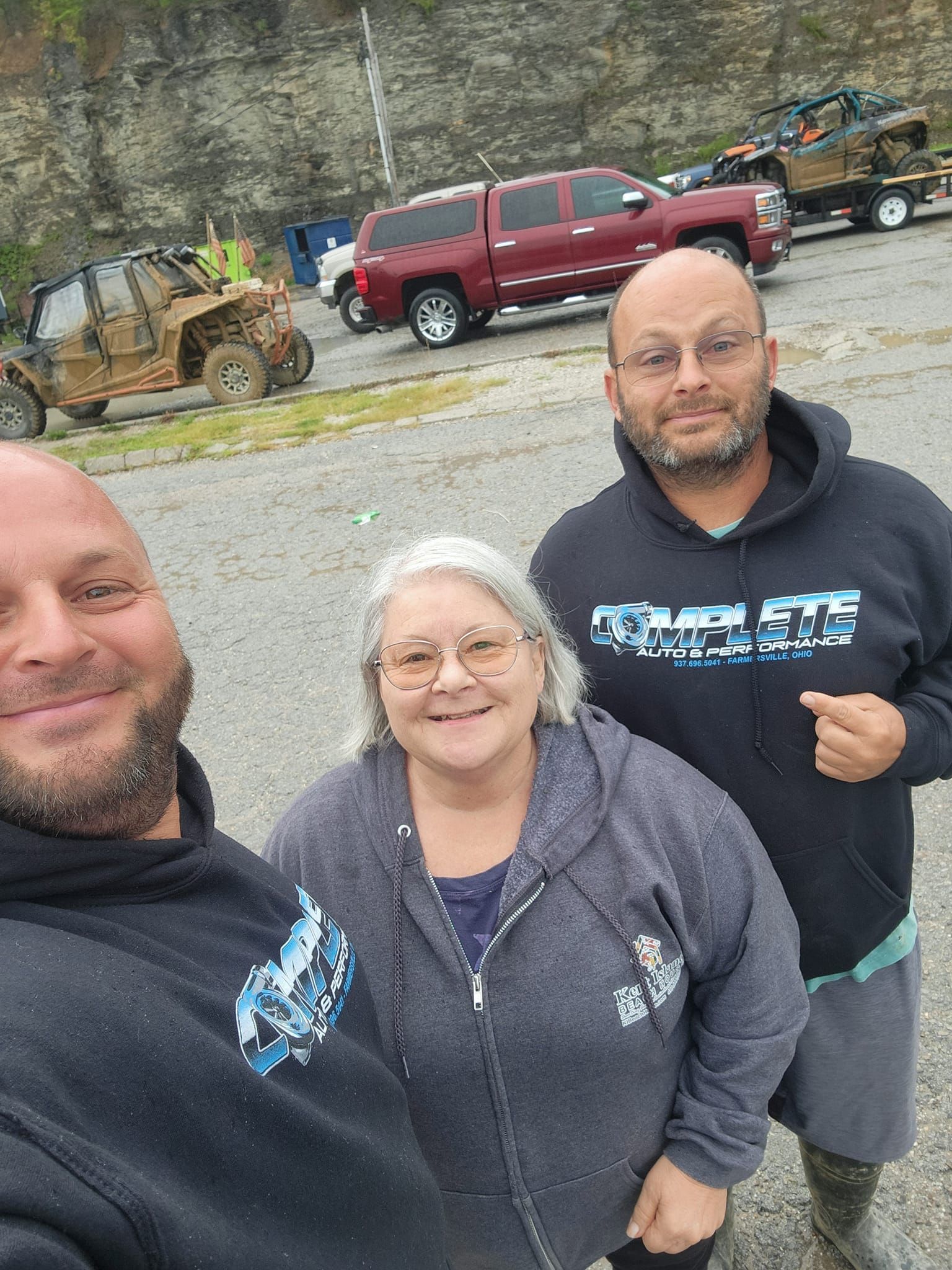 Three people smiling, posing for a photo. Vehicles and a trailer parked behind them outdoors.