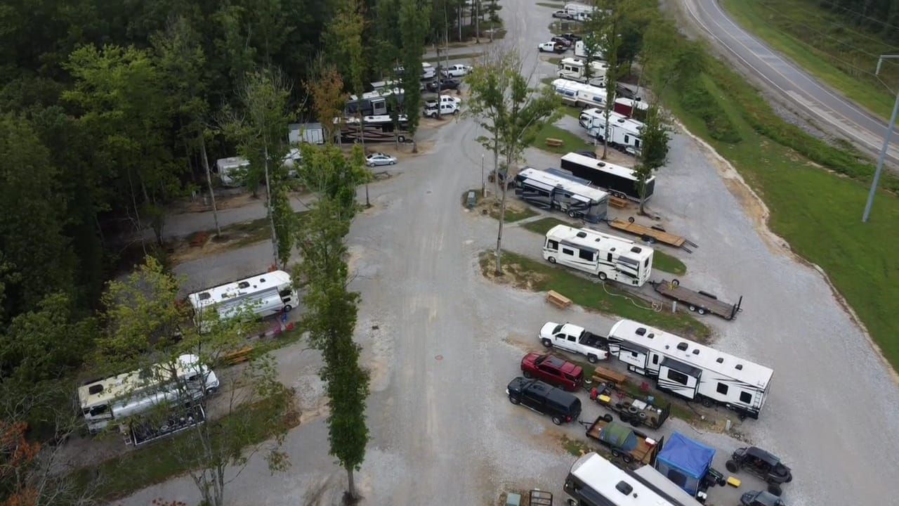 An aerial view of an RV park with vehicles parked on gravel, trees, and a road.