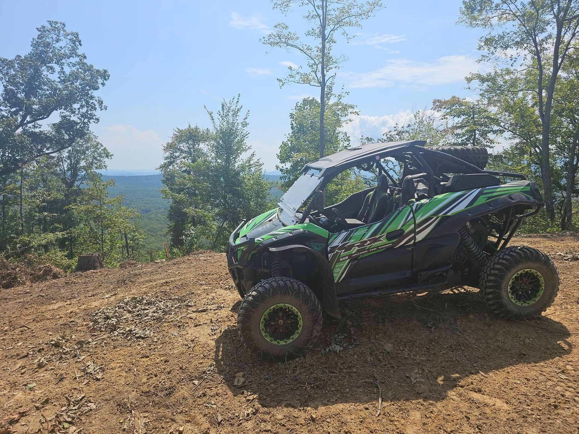 Green and black side-by-side ATV parked on a hillside with a view of trees and sky.