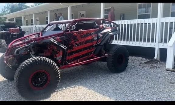 Red and black off-road vehicle with large tires parked on gravel, beside a white porch.