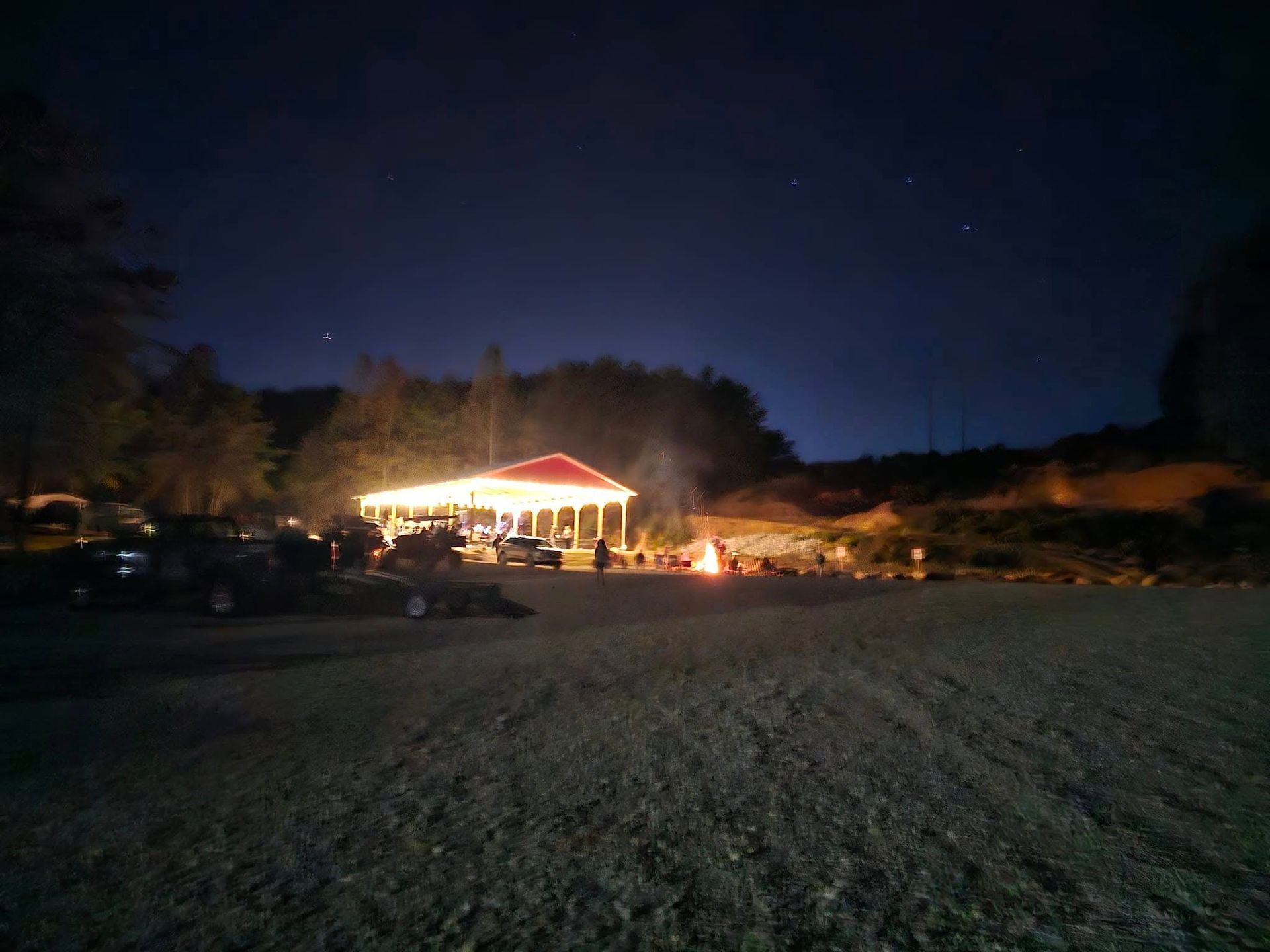 Night scene: lighted pavilion, people gathered, cars parked, dark sky with stars.