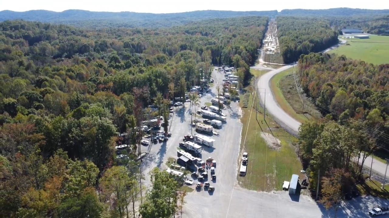 Aerial view of RVs and vehicles parked on gravel, surrounded by trees and a long highway.