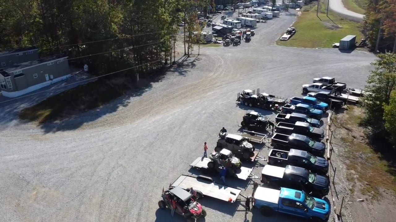 Vehicles parked in a gravel lot with buildings and trees in the background. Bright sunny day.
