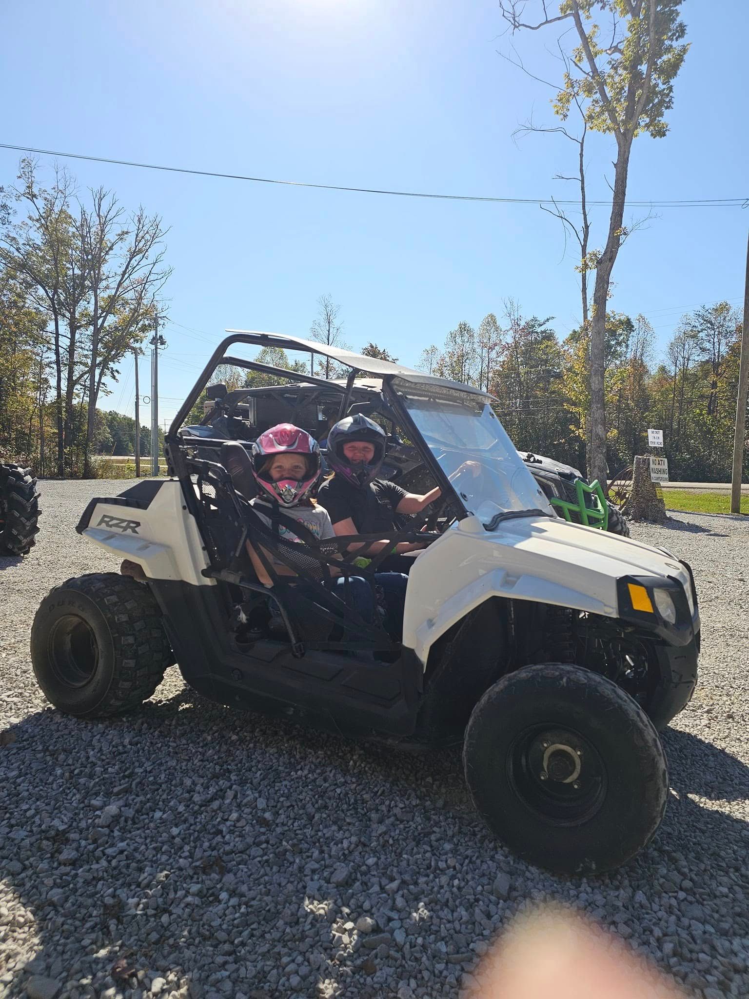 Two people in a white ATV with a roll cage, parked on gravel. Sunny outdoor setting.