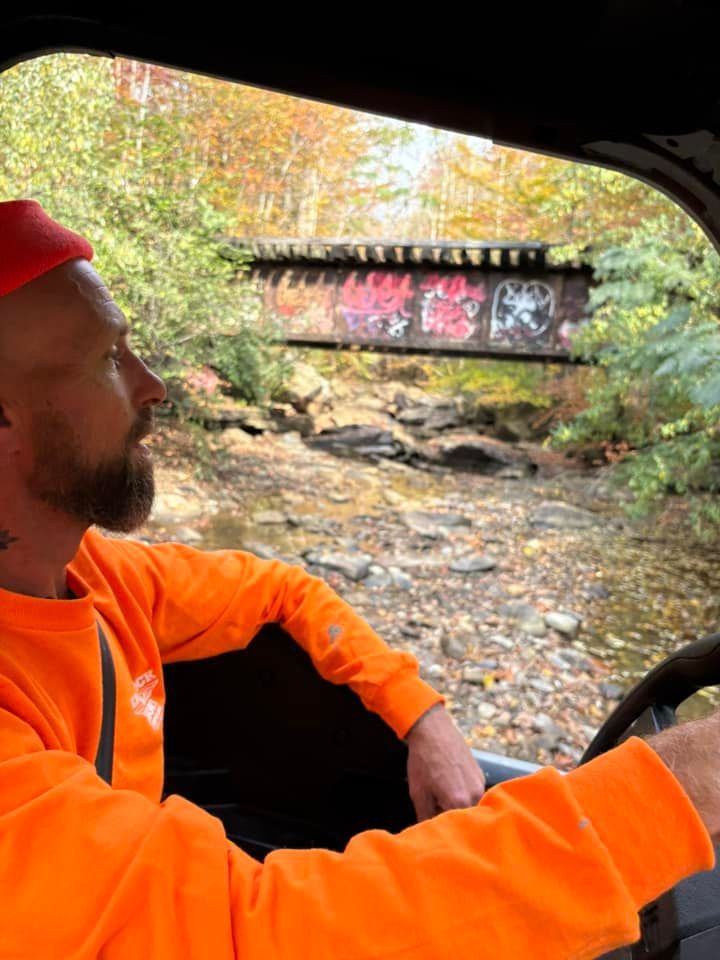 Man in orange driving an off-road vehicle toward a graffiti-covered bridge over a stream in a wooded area.