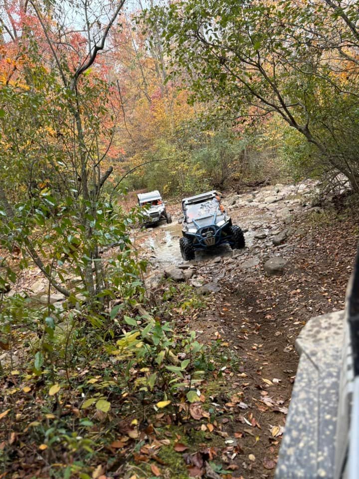 Two off-road vehicles drive through a rocky stream in a forest with fall foliage.
