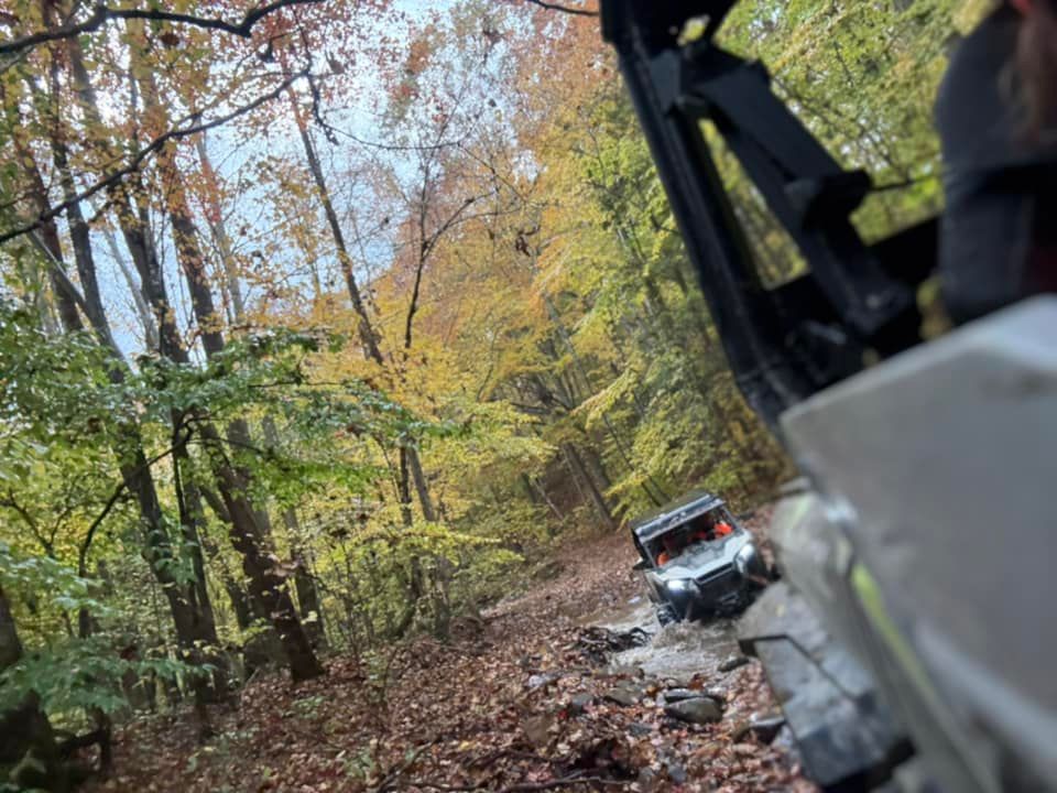 Side-by-side ATV traversing a muddy trail in a forest of autumn trees; partial view from another vehicle.