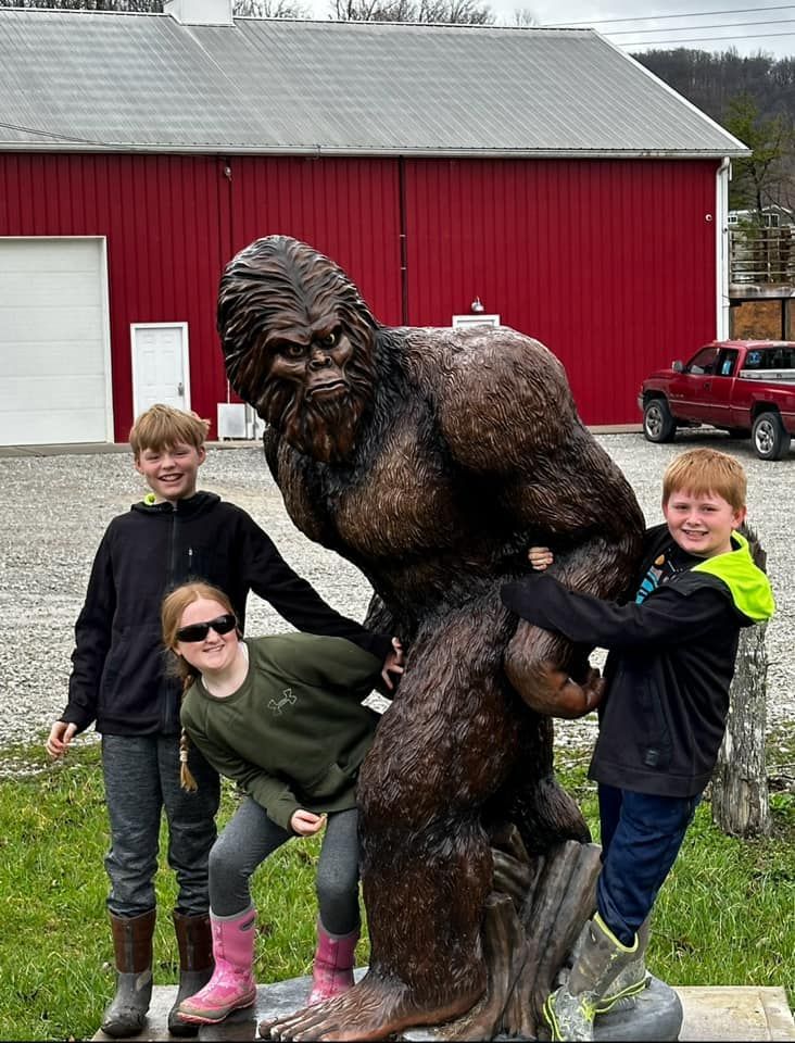 Three kids pose with a Bigfoot statue, laughing. A red barn and truck are in the background.