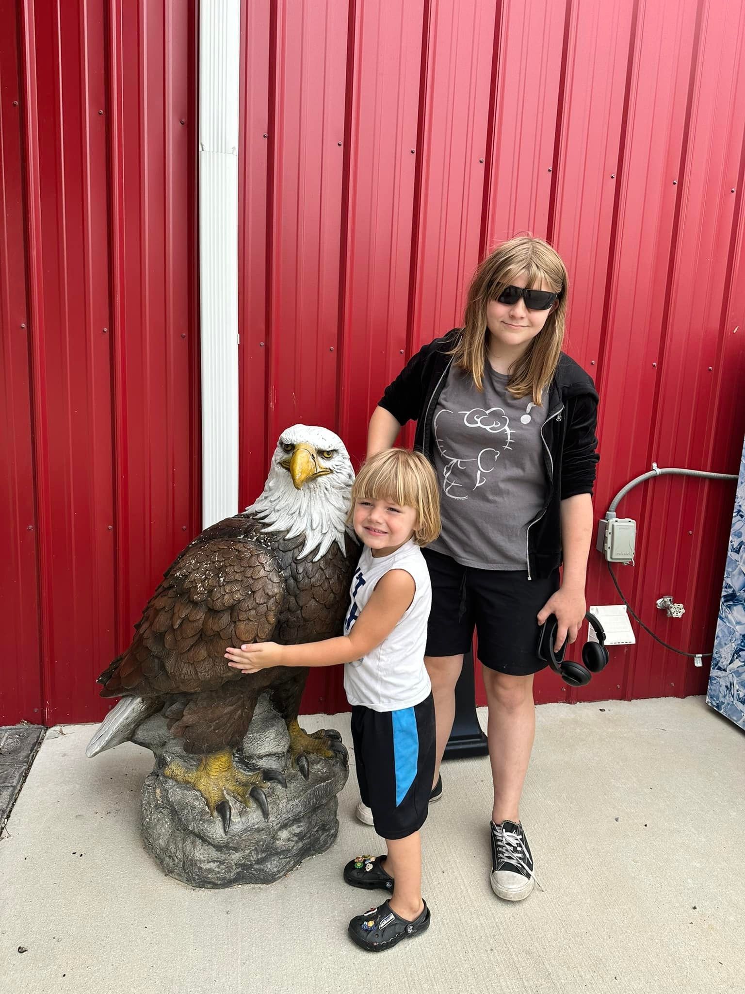 Two children pose with a large eagle statue in front of a red building.