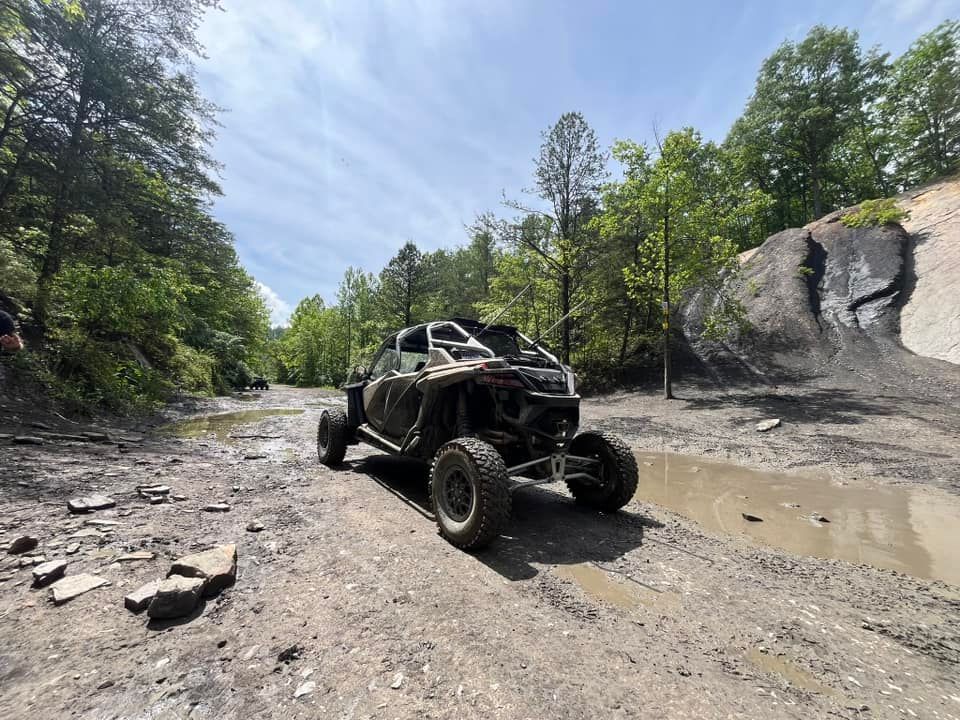 UTV driving on a muddy trail surrounded by trees. Blue sky overhead.