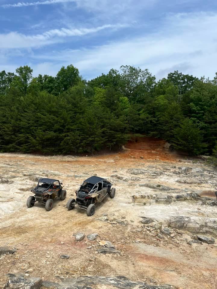 Two off-road vehicles parked on a rocky terrain with trees and a blue sky in the background.