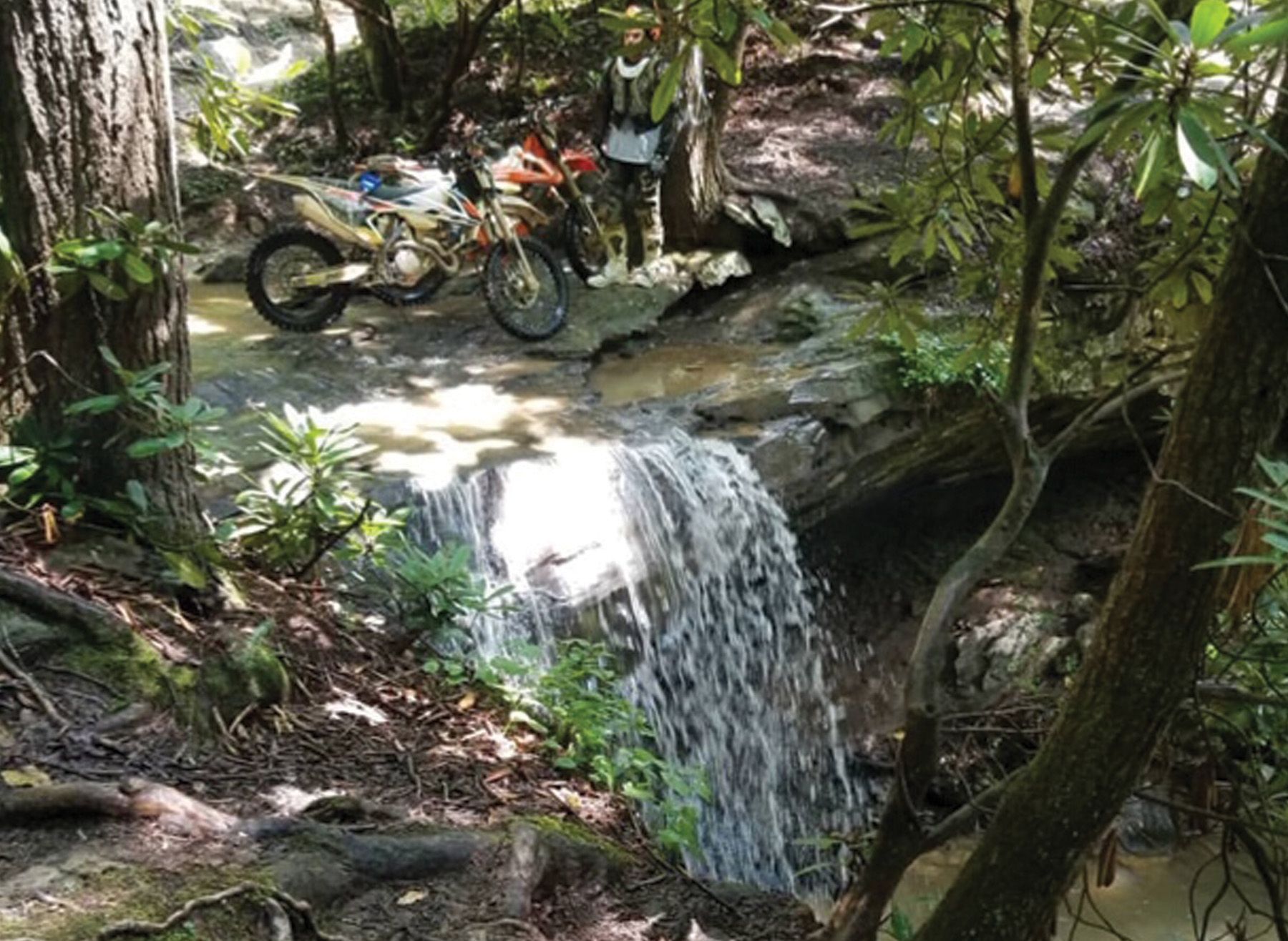 Motorcycle parked by a small waterfall in a forest. A person stands nearby, observing.