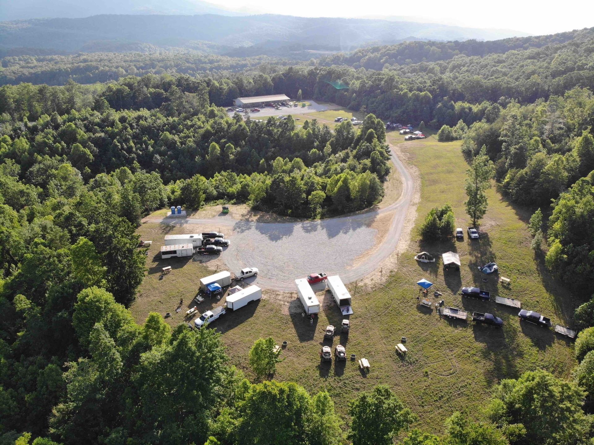 Aerial view of vehicles parked in a grassy clearing surrounded by trees, with a building in the background.