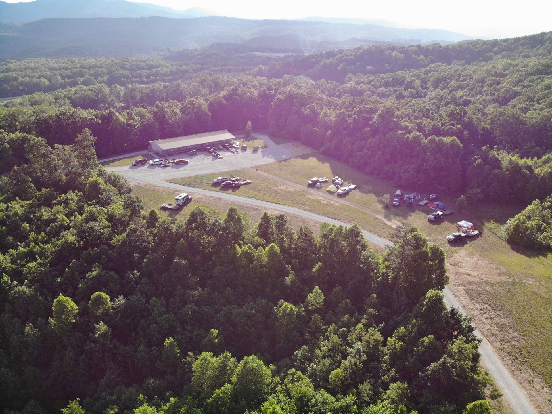 Aerial view of a clearing with a long building and vehicles surrounded by a dense forest, near a road.