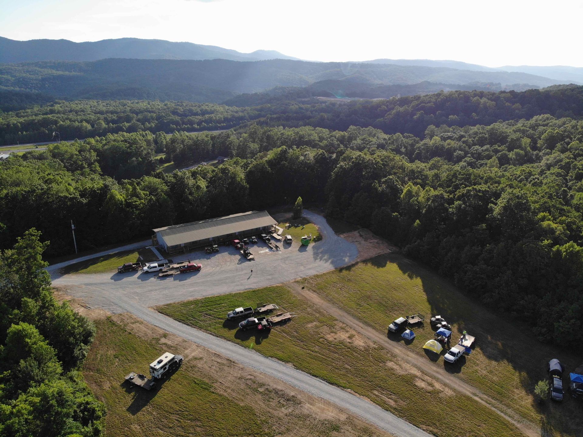 Aerial view of a building surrounded by trees and mountains with cars parked in a grassy area.