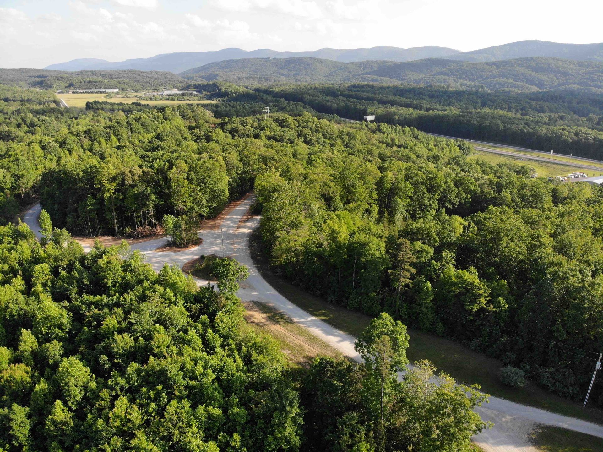Aerial view of a winding dirt road through lush green forest, with mountains in the distance under a blue sky.