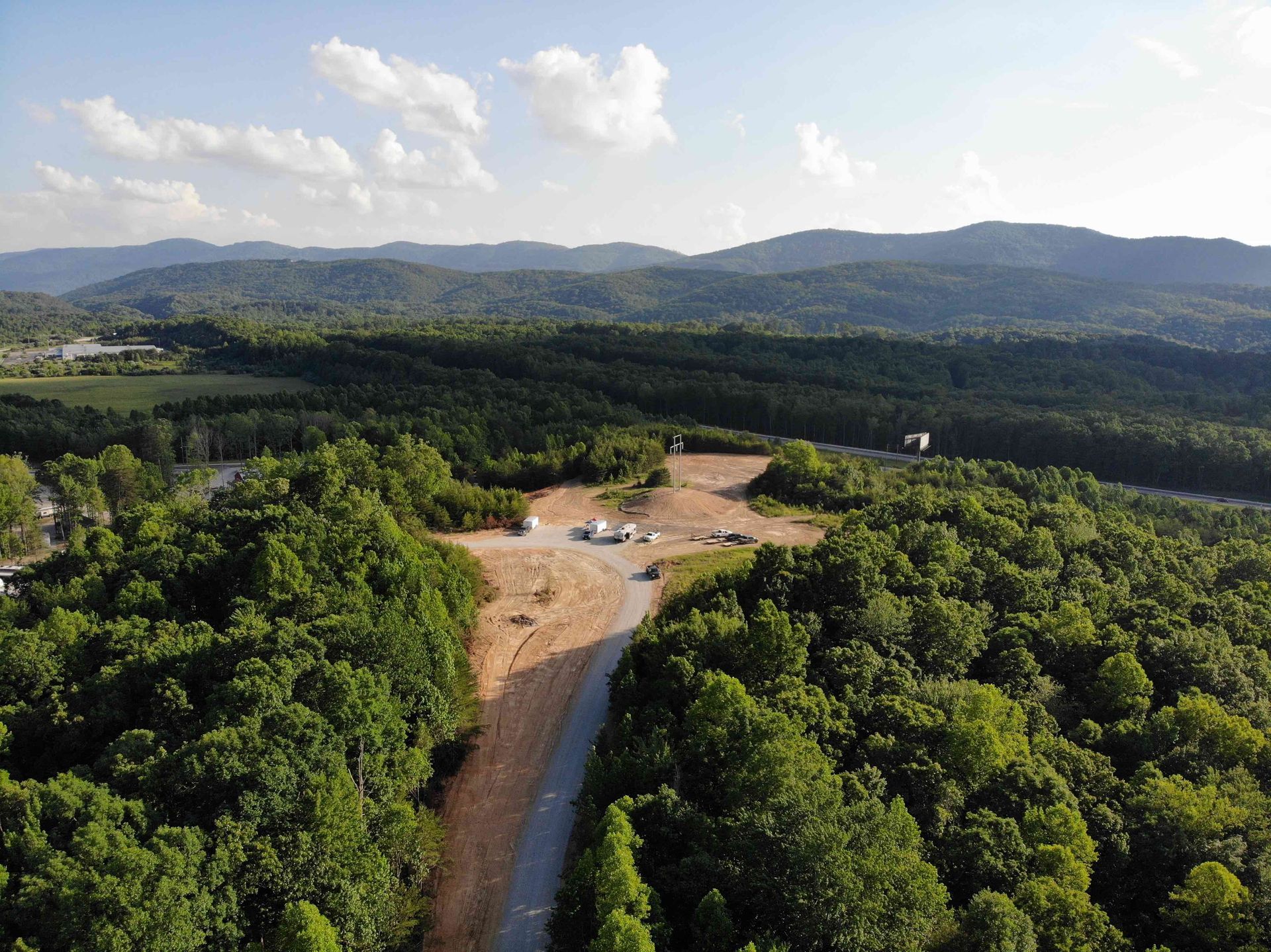 Aerial view of a clearing in a forest with a road leading to a construction site; green trees and distant hills.