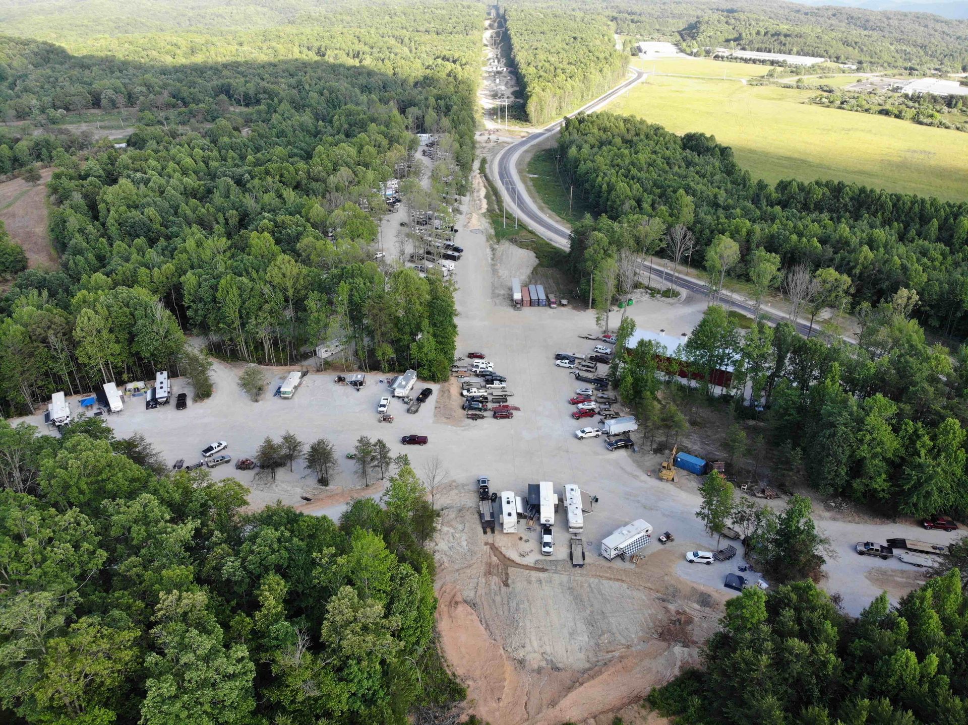 Aerial view of a road and adjacent gravel parking area with RVs, trees, and buildings.