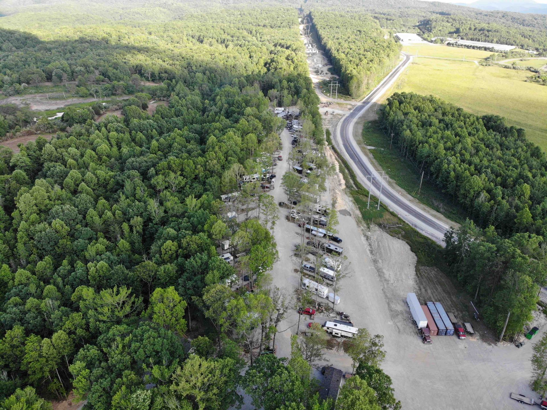 Aerial view: vehicles parked next to a road, bordered by a forest on one side and fields on the other.