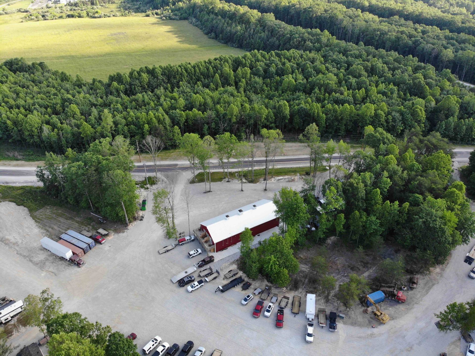 An aerial view of a red barn, gravel lot, vehicles, surrounded by trees and a road.