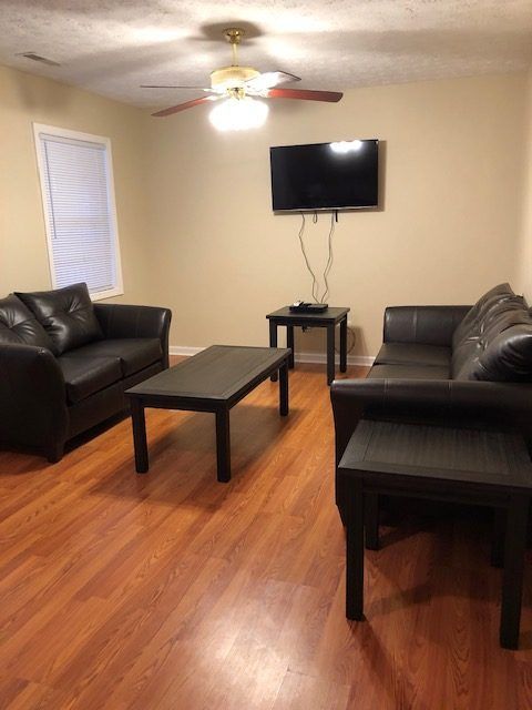 Living room with black furniture on wood floors; TV on wall, ceiling fan, window.