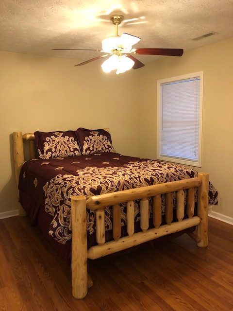 Bedroom with log-style bed frame, maroon bedding, and ceiling fan. Light-colored walls and wood floor.