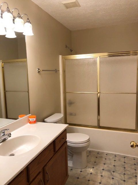 Bathroom with a toilet, vanity, and shower/tub combo. Gold trim, tan walls, and blue-gray patterned floor.