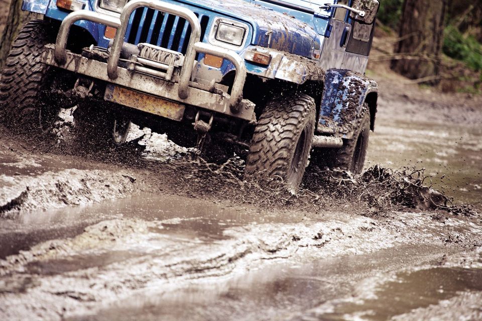Blue Jeep driving through muddy water, tires splashing, outdoor setting.