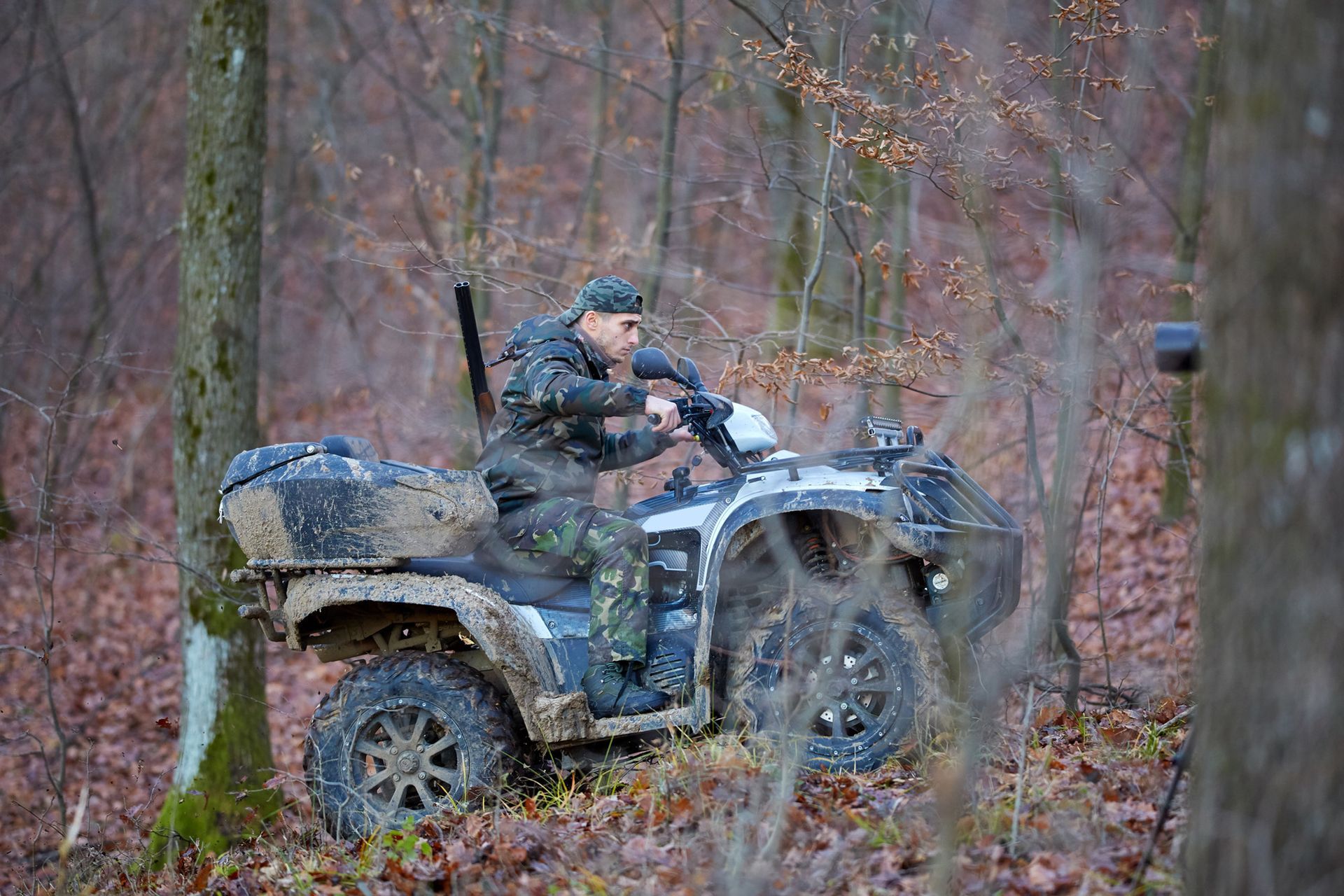 Man on ATV in camouflage clothing drives through a muddy forest.