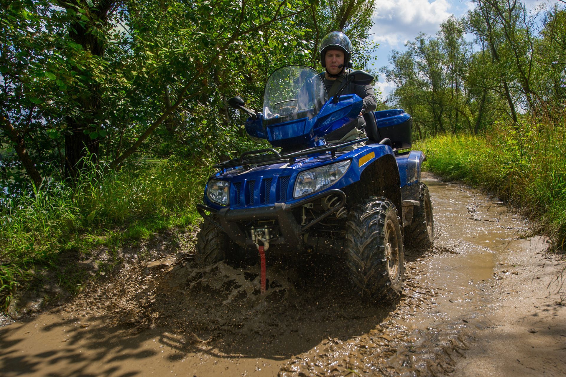 Person on a blue ATV driving through a muddy puddle on a dirt path, trees on either side.