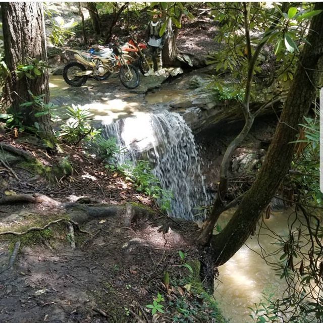 Dirt bike next to a small waterfall in a forest. A person stands nearby, looking towards the bike.