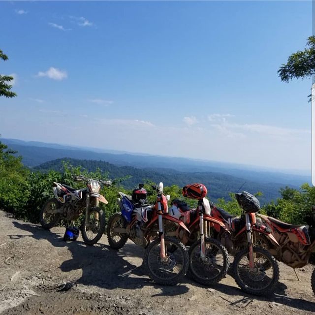 Dirt bikes parked on a hillside overlooking a forested valley under a blue sky.