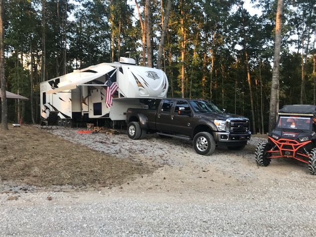 A fifth-wheel camper hitched to a dark pickup truck with an ATV parked nearby at a campsite.