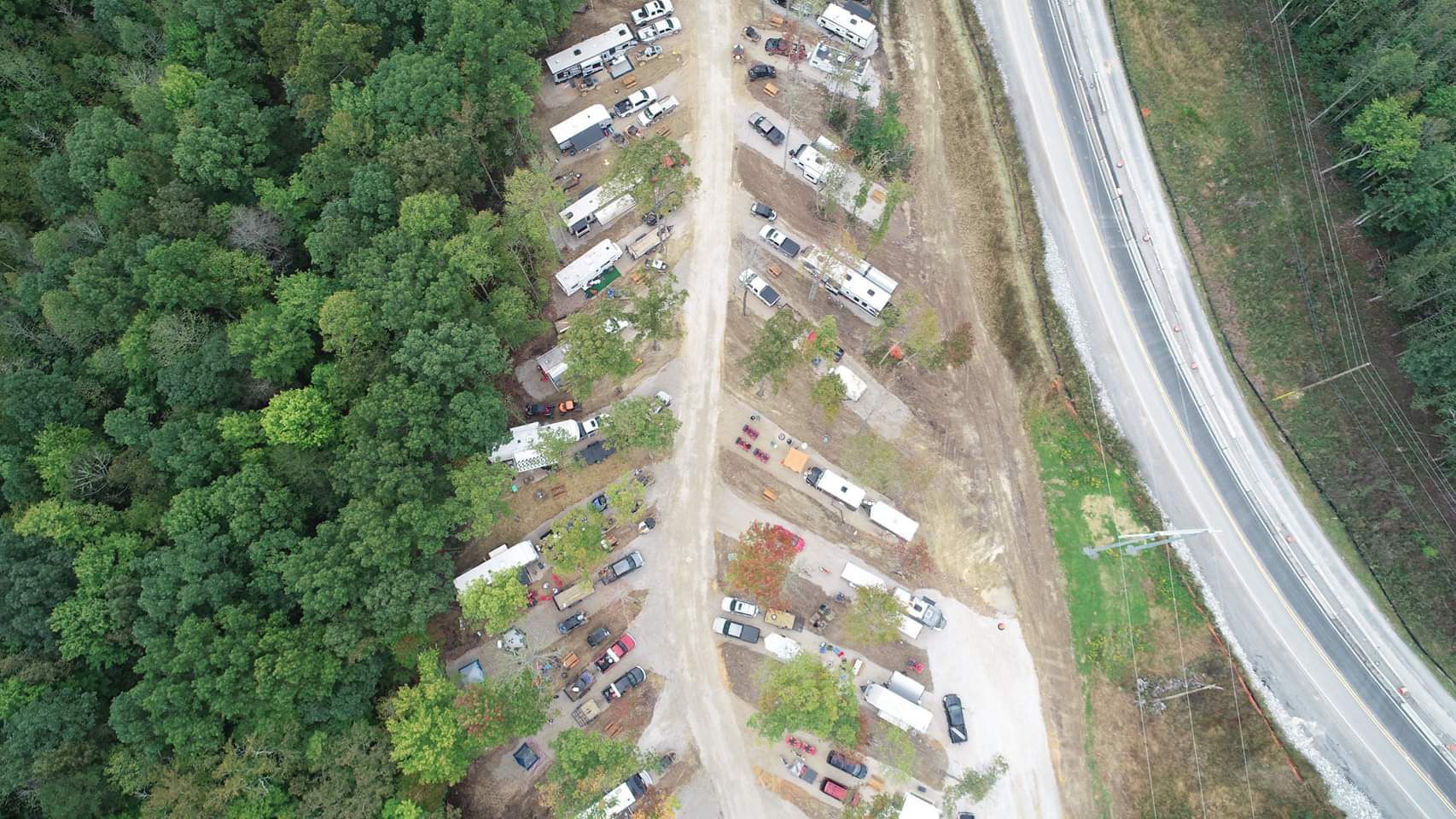 Aerial view of RV park with vehicles parked along a central road, next to a highway and forest.