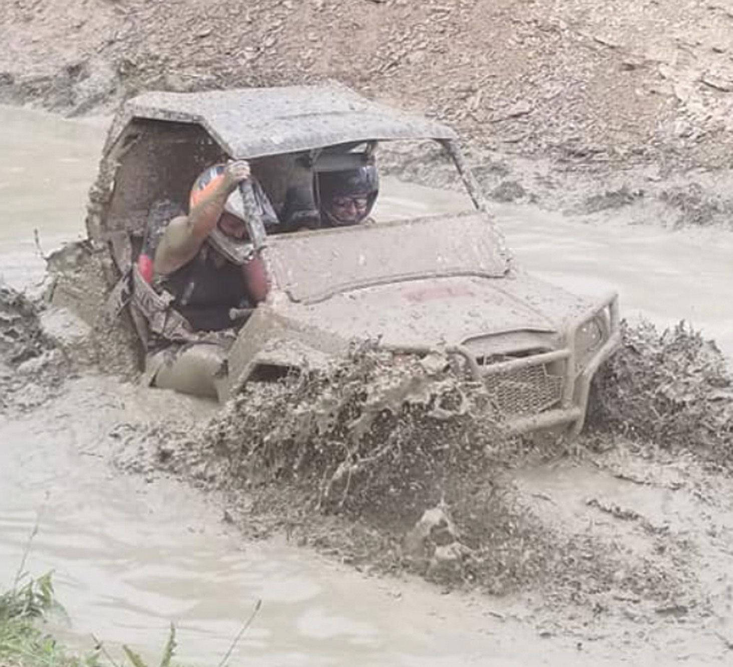 Mud-covered ATV splashing through muddy water, two people inside, driver raising hand, outdoor setting.