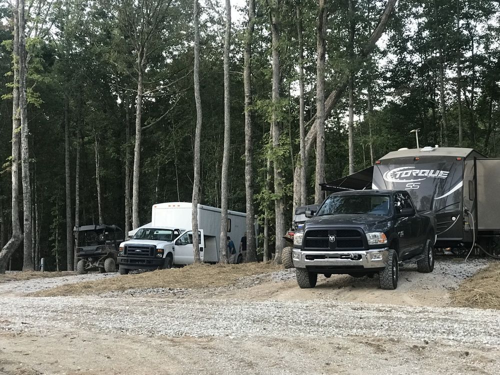 Vehicles parked at campsite with RV. Black truck, white trailer, ATV and tall trees in background.