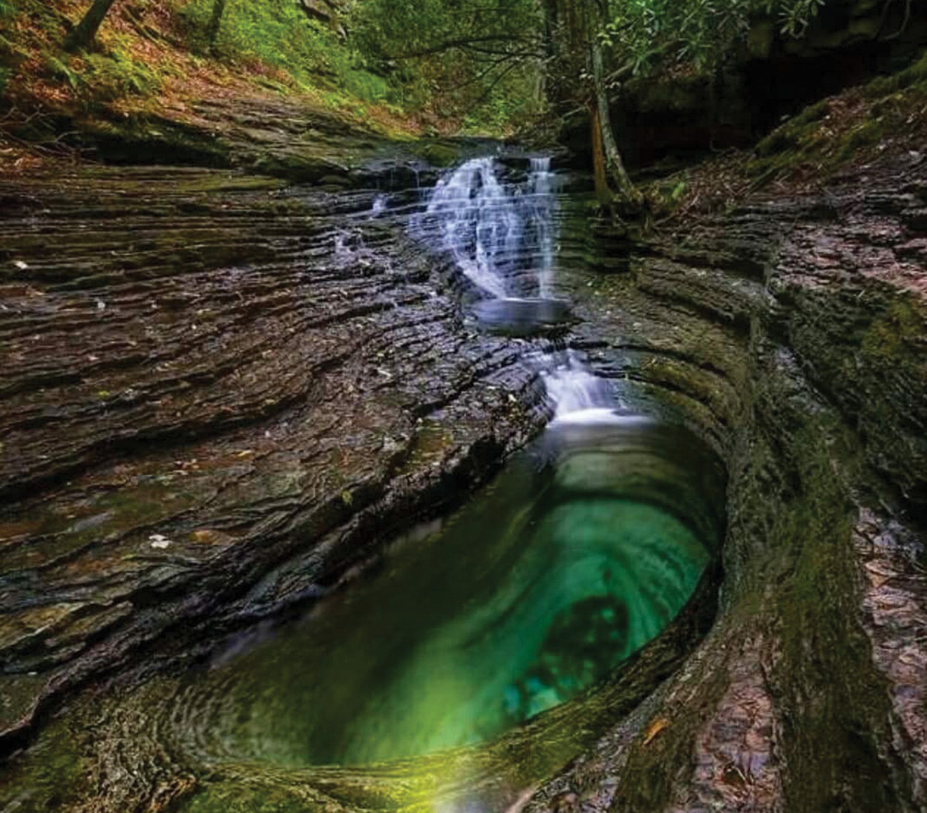 Waterfall cascading into a turquoise pool carved in rock. Dark, layered rock surrounds the water.