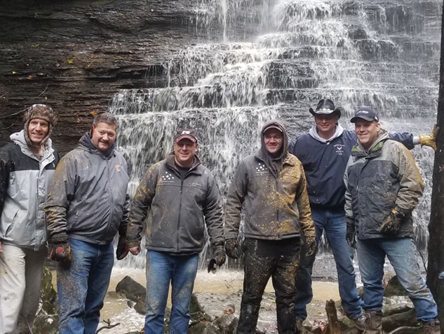 Six men pose in front of a waterfall, covered in mud. Some wear hats and jackets.