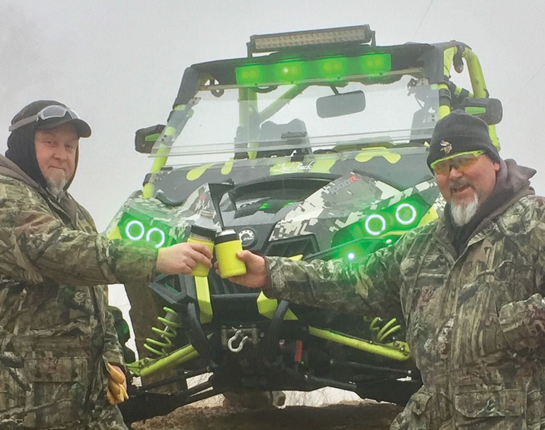 Two men in camouflage toasting with mugs in front of a neon green and camo UTV.