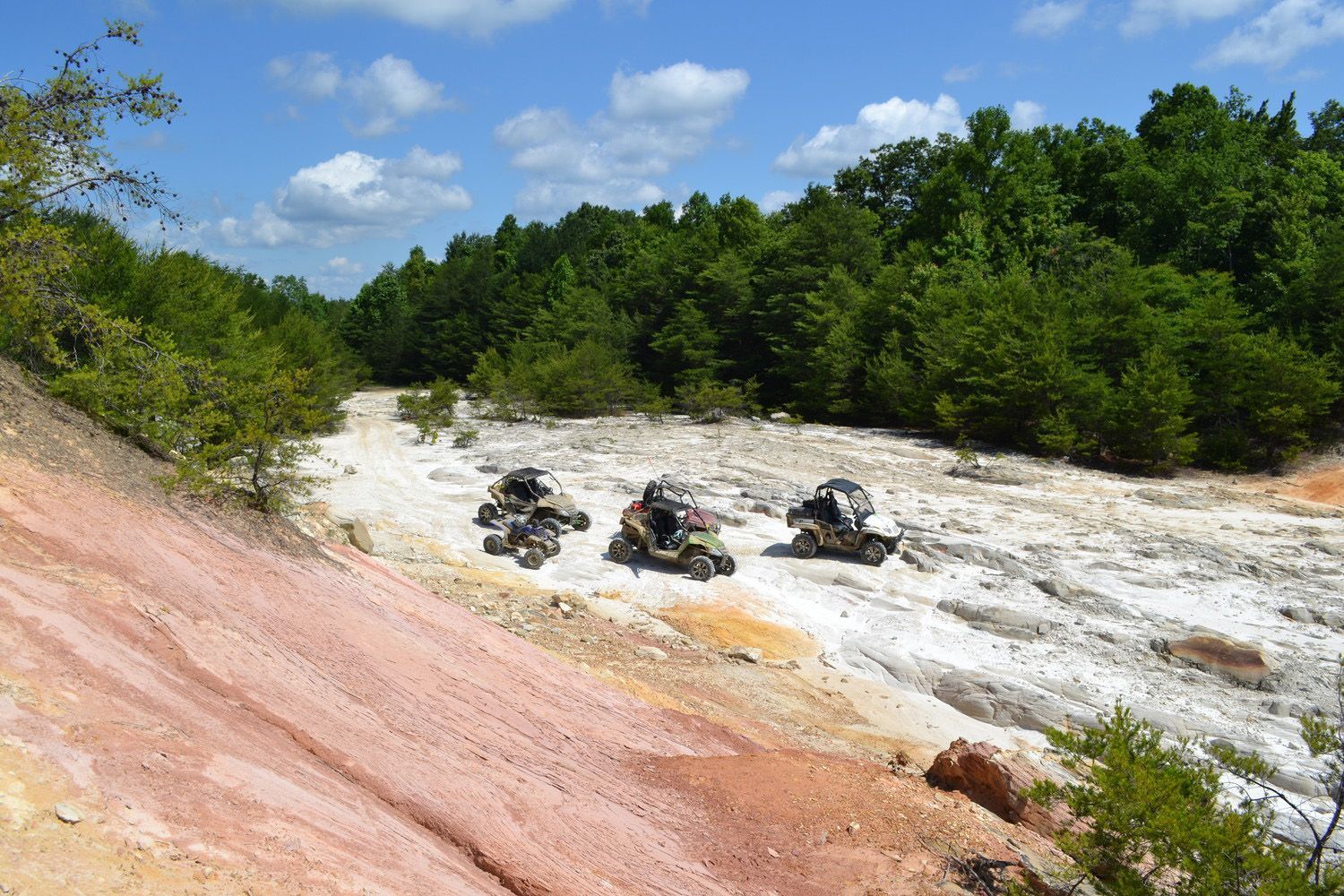 Three ATVs parked on a white and red dirt trail in a forest clearing on a sunny day.