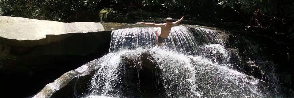 A person with arms outstretched stands on a waterfall, surrounded by lush green trees.