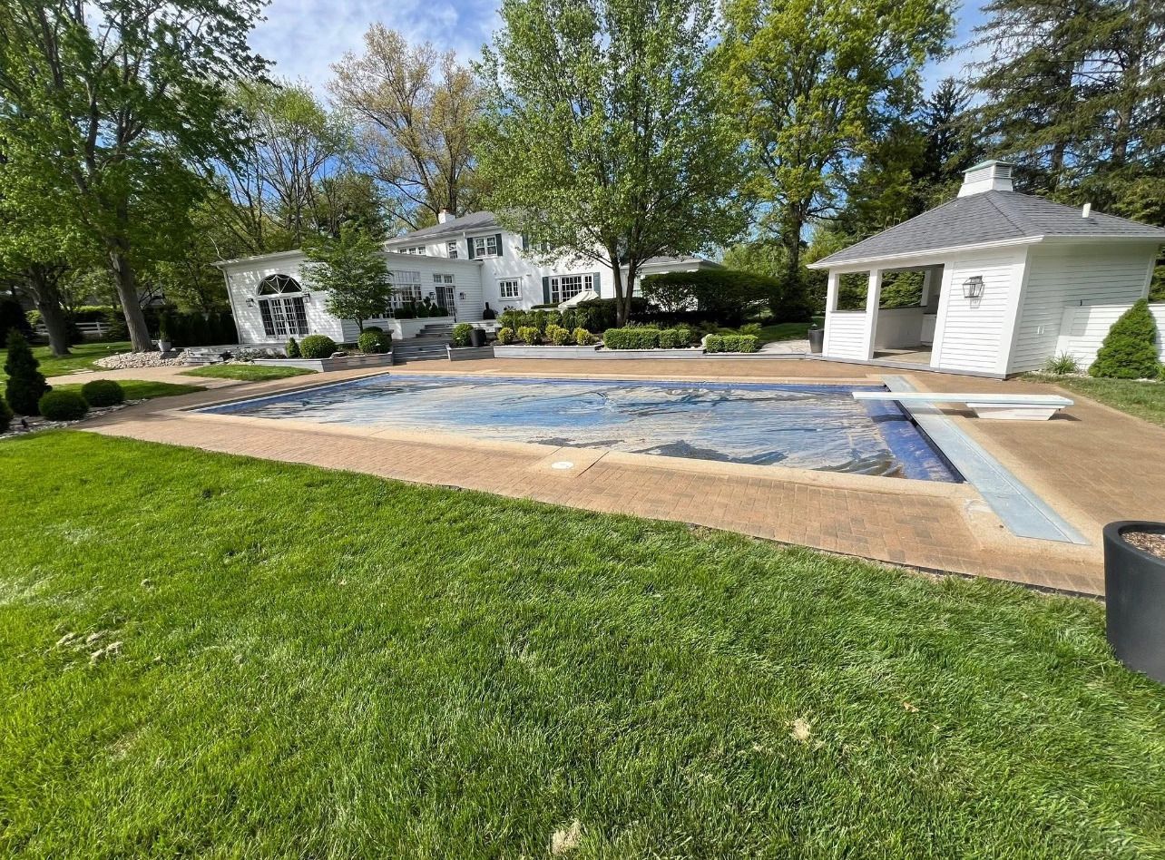 Backyard with a covered rectangular pool, green lawn, and white pool house under trees