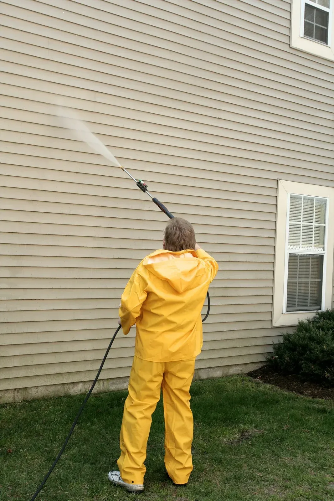 A man in a yellow raincoat is cleaning the side of a house with a high pressure washer.